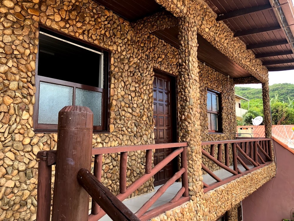The exterior of the stone apartment showcases a unique textured surface with natural stones arranged decoratively. A wooden railing borders the covered porch, while large windows and a double wooden door provide access to the interior. The surrounding landscape hints at lush greenery.