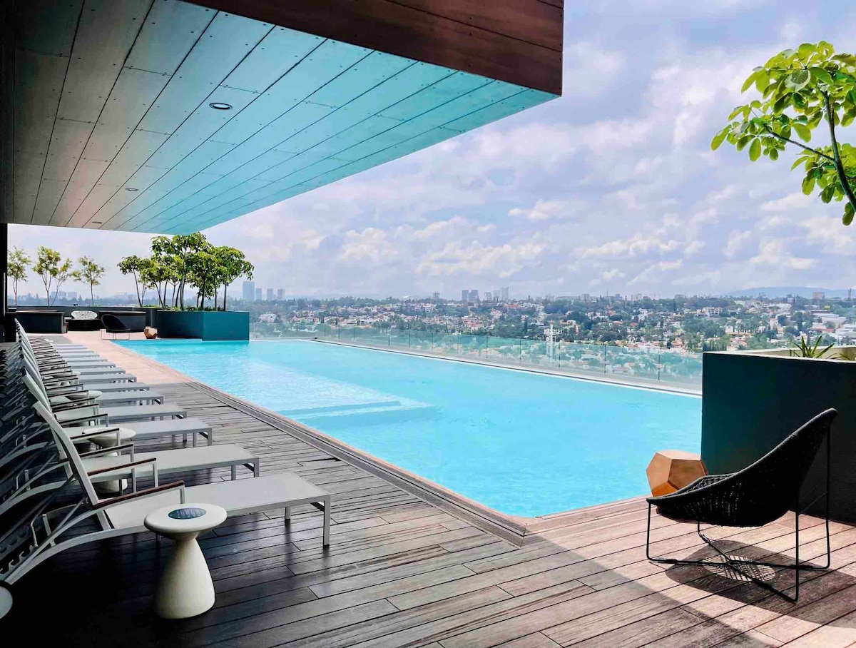 An infinity pool overlooks the city, surrounded by modern lounge chairs positioned on a wooden deck. Lush greenery is visible in the background, enhancing the serene atmosphere. The skyline is framed under a partly cloudy sky, offering a sense of openness and tranquility.