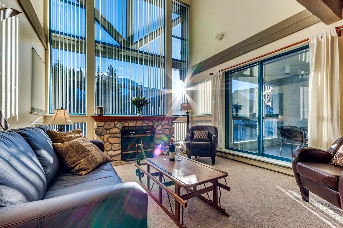 A spacious living area is highlighted by large windows, allowing natural light to fill the room. A stone fireplace serves as a focal point, complemented by comfortable seating. The room features a mix of textures, from soft carpeting to sleek furnishings, creating a cozy yet sophisticated atmosphere.