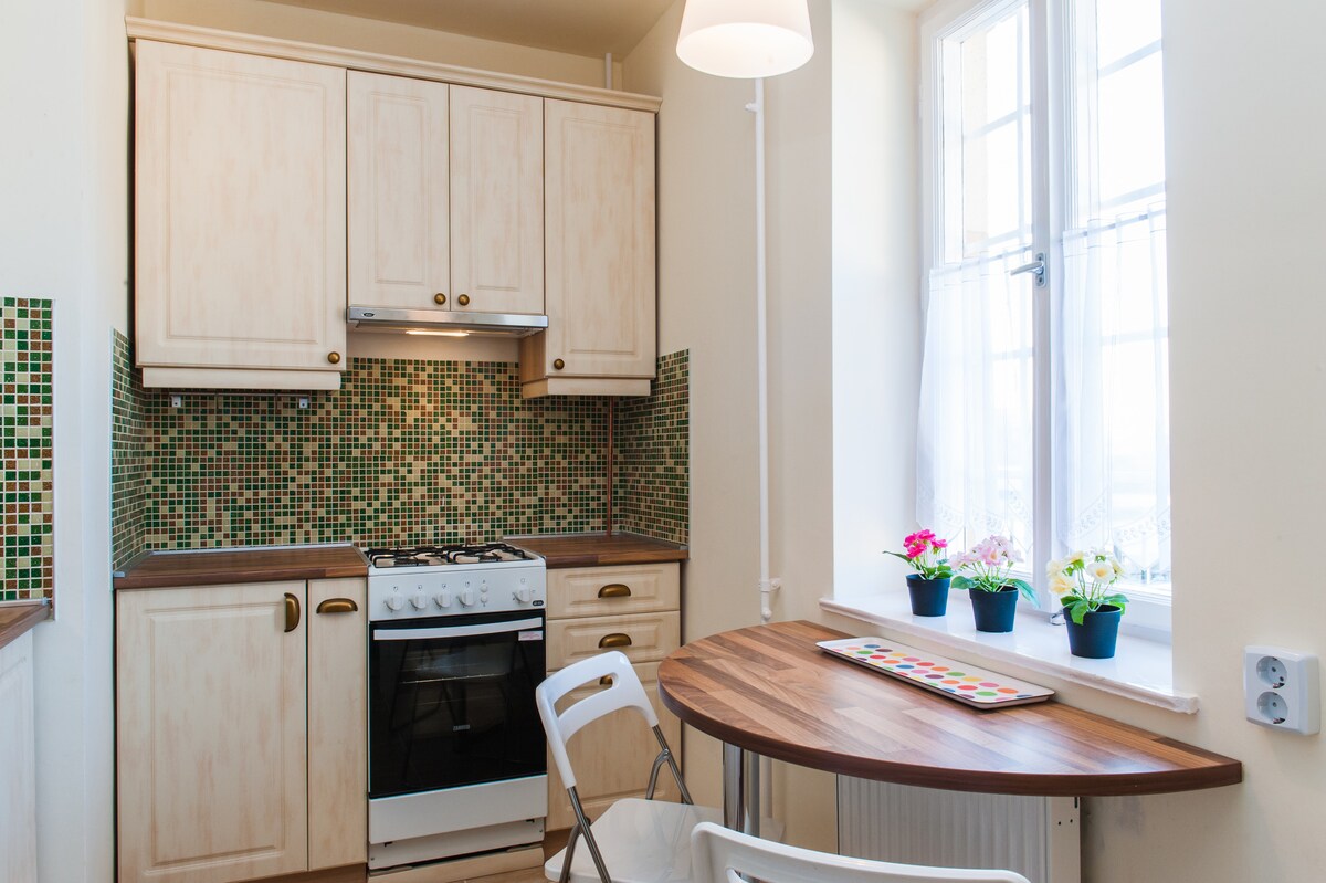 The kitchen features light-colored cabinetry complemented by a mosaic tile backsplash. A well-organized countertop includes a stove and a small dining table with a colorful centerpiece. Natural light enters through two windows adorned with sheer curtains, enhancing the pleasant atmosphere.