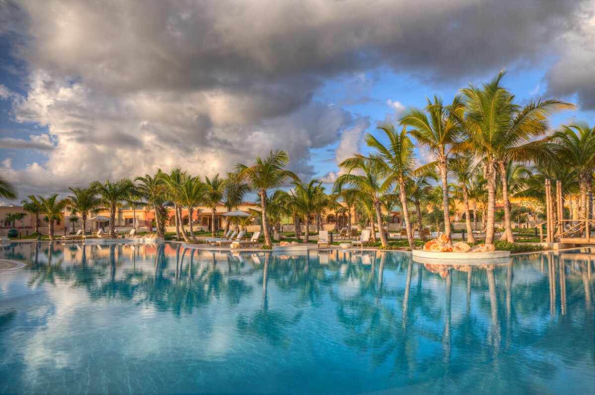 A large pool is surrounded by palm trees and lounge chairs, creating a serene environment. The water reflects the passing clouds and surrounding landscape, contributing to a tranquil atmosphere. Several cabanas are visible along the pool's edge, providing shaded seating options.