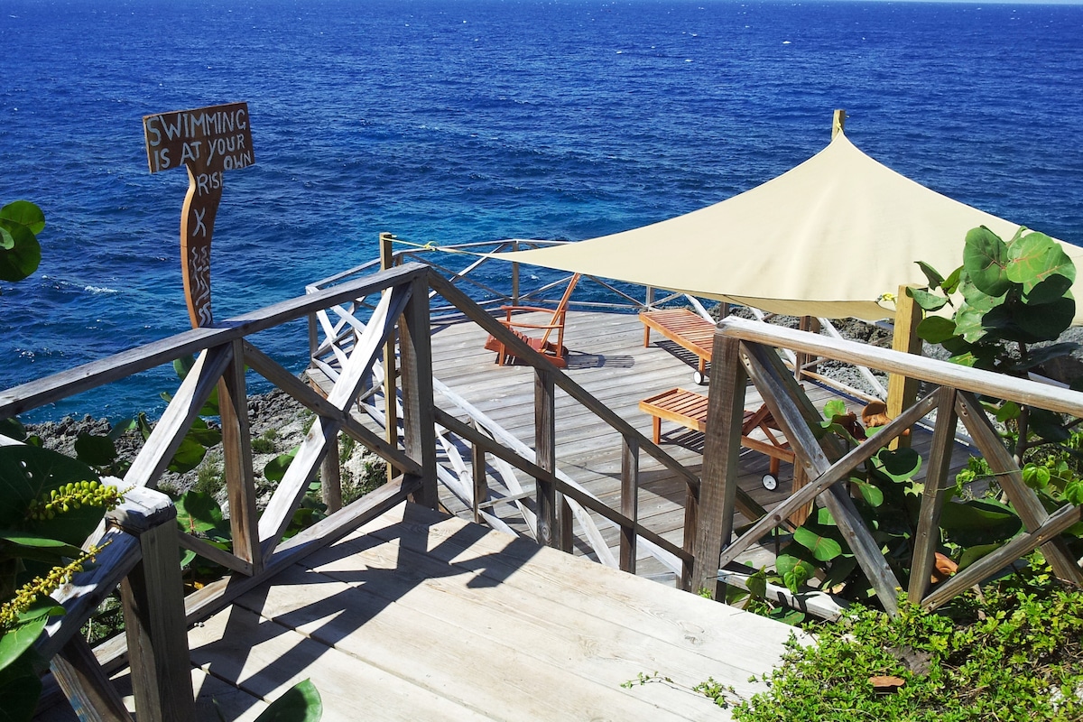 A wooden deck extends toward the ocean, accessed by a staircase with side railings. A large sun shade is positioned above two wooden loungers, providing a shaded area. A sign warns that swimming is at one's own risk, framed by lush green foliage.