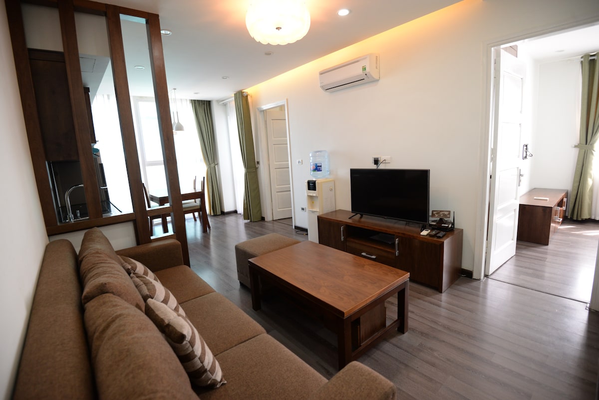 A comfortable living area is furnished with a brown sectional sofa and a wooden coffee table. A flat-screen television is positioned on a console, with an air conditioning unit mounted above. Natural light enters through large windows, and a dining table for four is visible in the background.