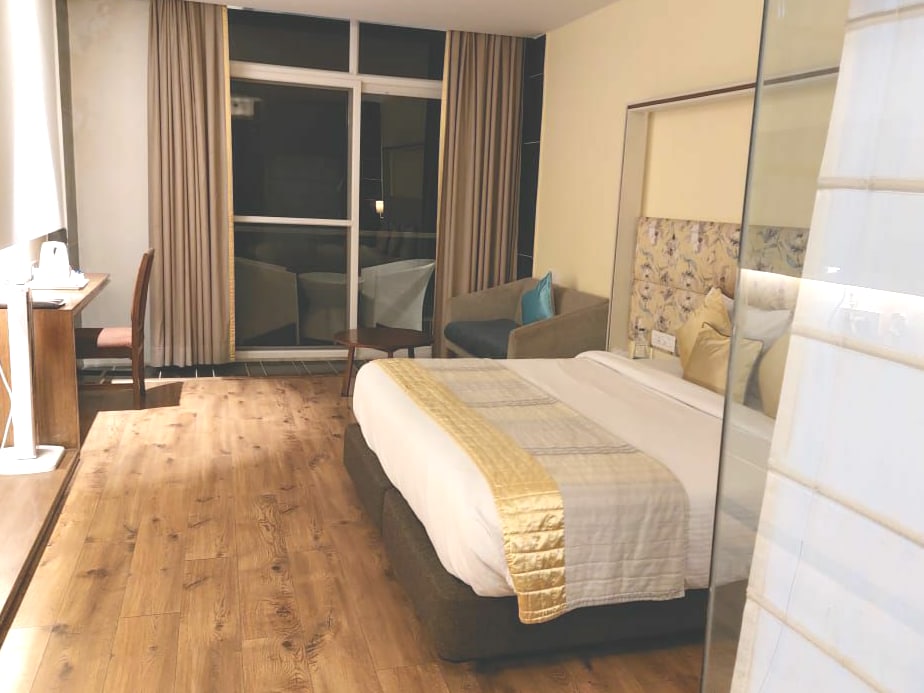 A spacious room features a large bed with a light-colored coverlet. A comfortable seating area with a sofa is present, and natural light enters through large windows adorned with sheer curtains. A work desk is positioned against the wall, complementing the wooden flooring.