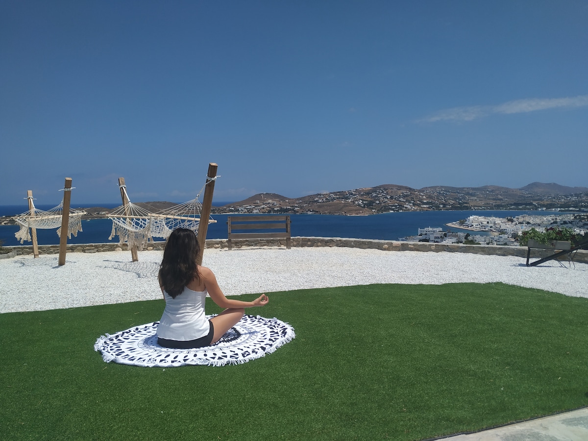 A tranquil outdoor space features a person meditating on a patterned mat. In the background, a stunning view of the Paroikia gulf is visible, framed by rolling hills and a clear blue sky. Two hammocks are positioned nearby on a manicured lawn.
