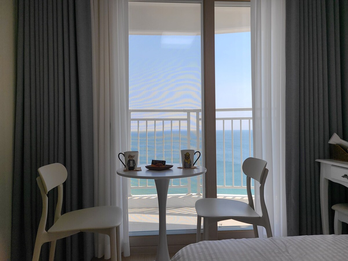 A small round table is set by two white chairs, facing a large window with sheer curtains, allowing a view of the ocean. Sunlight filters through, illuminating the table, which holds two cups and a small plate. The calm blue waters are visible beyond the railing.