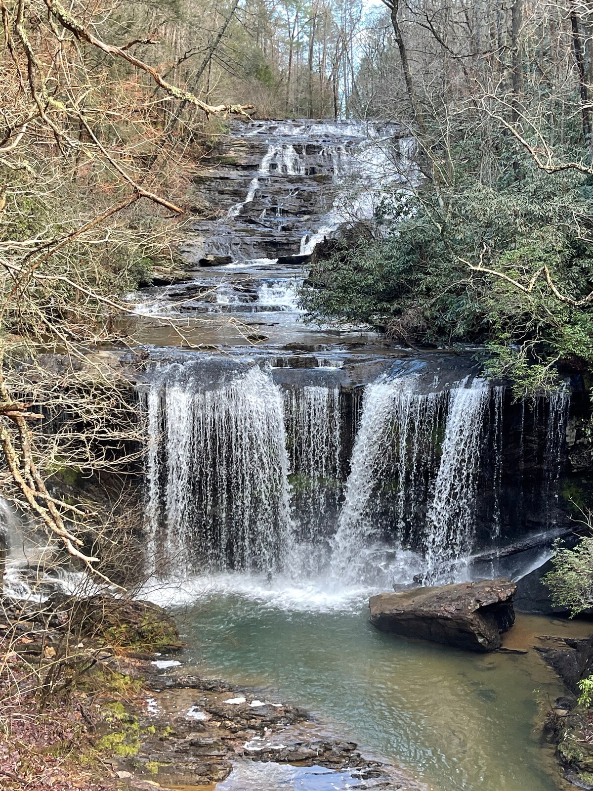 A serene waterfall cascades down rocky steps, creating a tranquil pool at its base. Surrounded by lush greenery and large boulders, the scene embodies a peaceful natural habitat, with water flowing gently over the rocks.