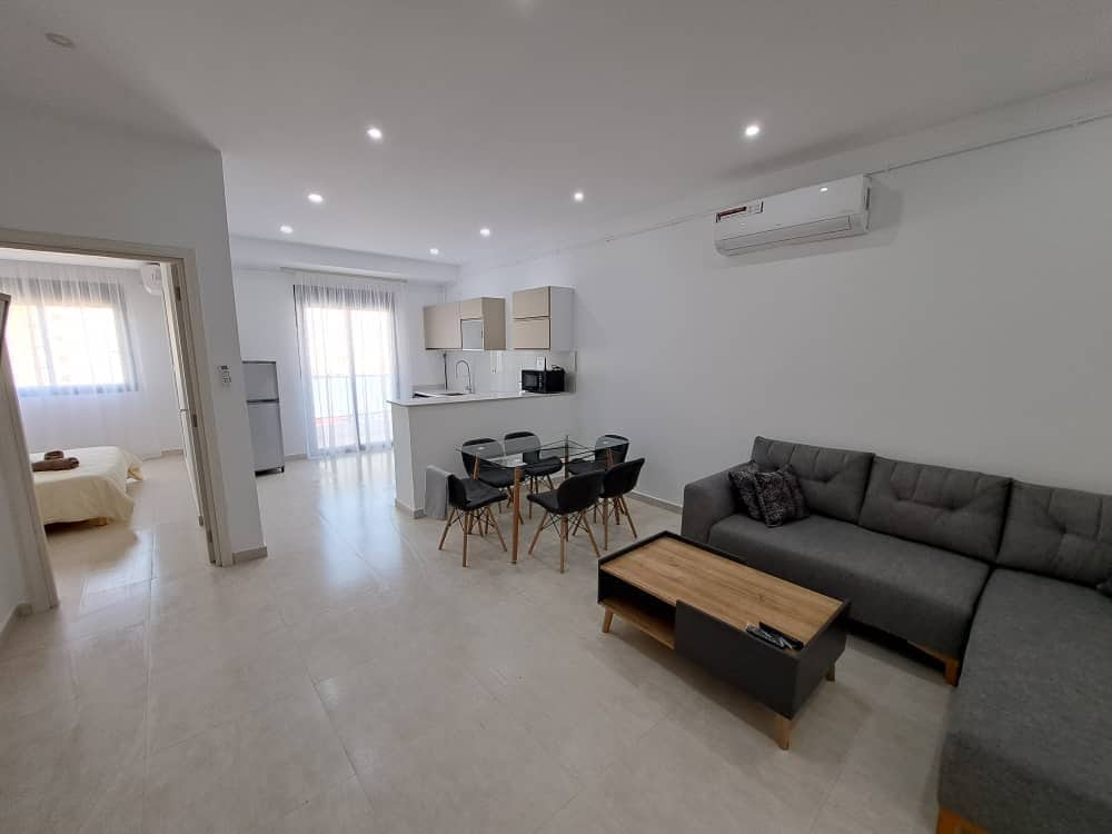 A spacious living area features a gray sectional sofa and a wooden coffee table. A glass dining table is surrounded by four chairs. The open kitchen is equipped with modern appliances, and large windows provide natural light, enhancing the airy atmosphere.