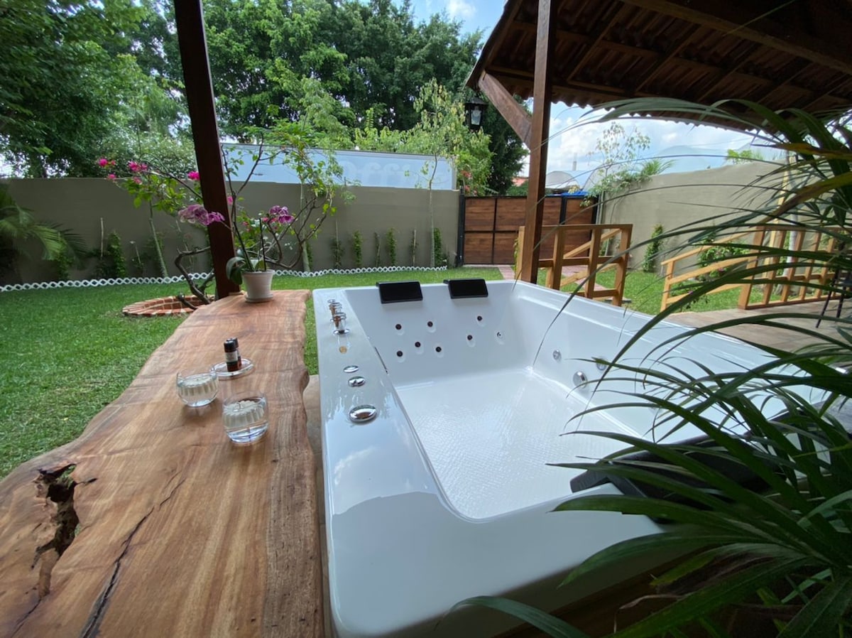 A private jacuzzi is situated under a wooden shelter, surrounded by greenery. A wooden table is seen in the foreground, adorned with small glasses and a decorative item. The setting offers a serene atmosphere, enhanced by the lush landscaping in the background.