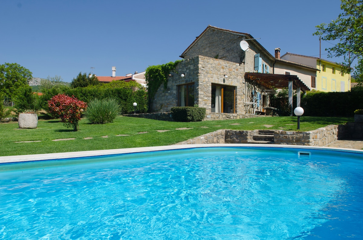 A semi-detached villa made of stone is set against a clear blue sky. The villa features large windows and is surrounded by well-maintained greenery. The inviting pool reflects the sunlight, while a patio area with outdoor seating is visible, enhancing the outdoor space.