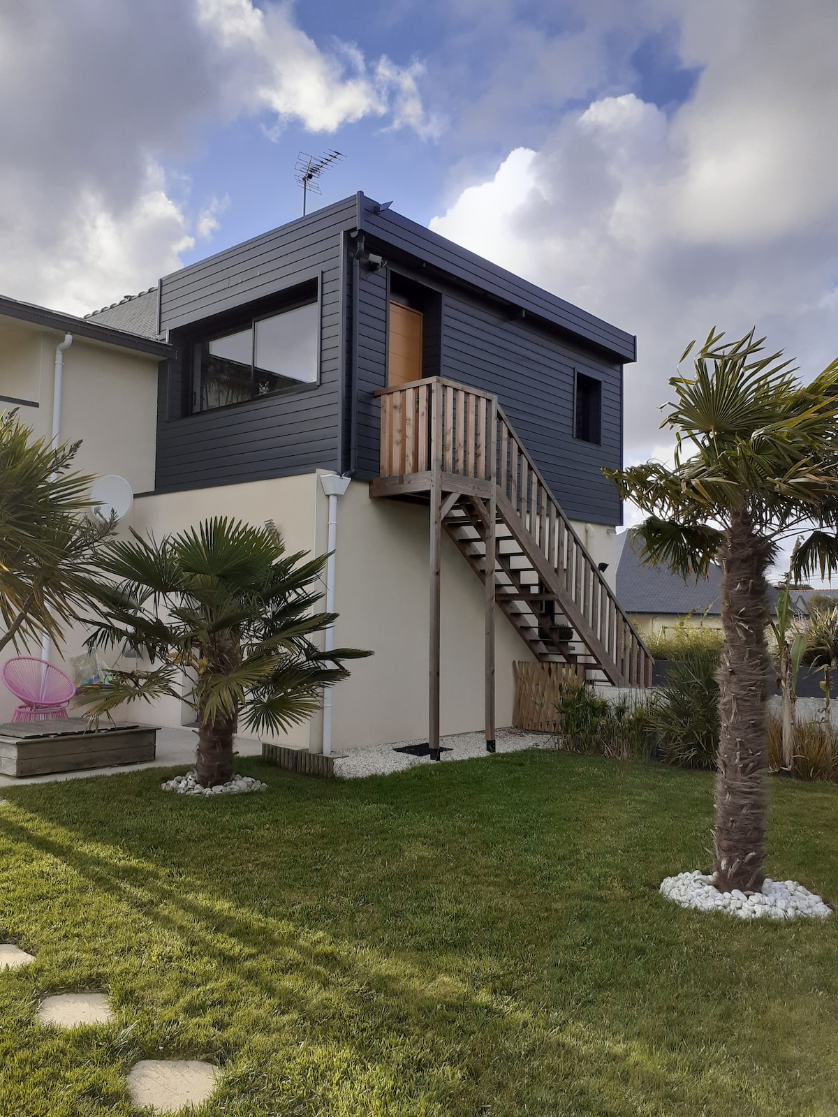 An exterior view of the house features a modern upper level with a wooden staircase leading to the entrance. Lush palm trees and a manicured lawn surround the base, contributing to a serene outdoor environment. The sky is partly cloudy, enhancing the tranquil setting.