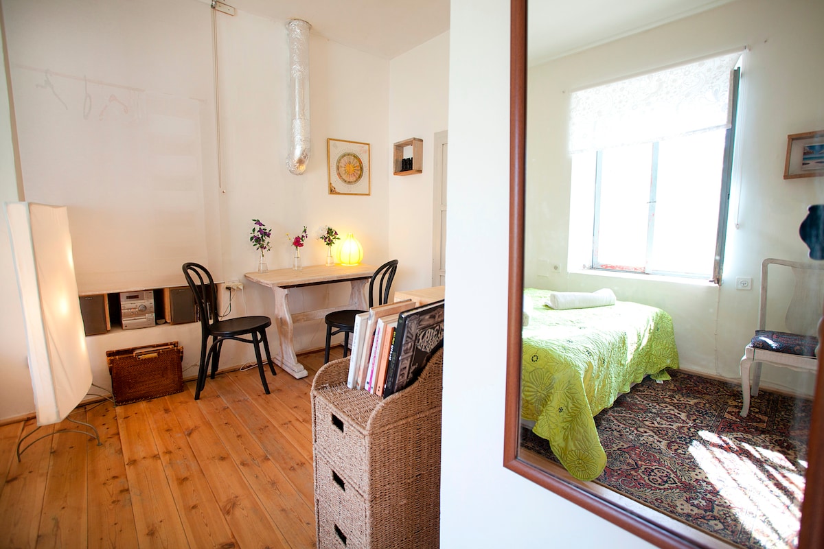 The studio apartment features a wooden floor and a cozy layout. A table with chairs is positioned near a window, creating a bright work area. A spacious bed is adorned with a green coverlet, and a woven storage unit holds books, adding to the welcoming ambiance.