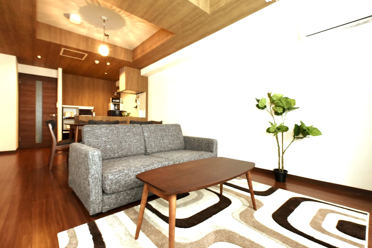 The living area features a cozy gray sofa paired with a low wooden coffee table, set on a patterned rug. A tall potted plant adds a touch of greenery, while warm wood tones and recessed lighting create a subtle ambiance.