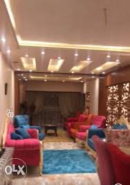 A bright living area is featured, furnished with cozy seating in various colors, including blue and pink sofas. A low coffee table is centered on a soft blue rug, while warm lighting and elegant decor enhance the ambiance.