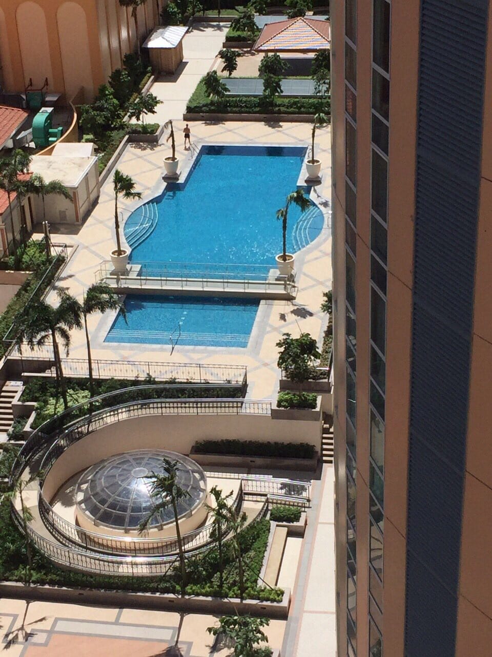 A spacious outdoor pool area is visible, surrounded by lush greenery and well-maintained landscaping. The pool features a clear, inviting blue surface, with lounge areas and decorative elements like palm trees. A domed structure adds architectural interest, enhancing the overall ambiance of the space.