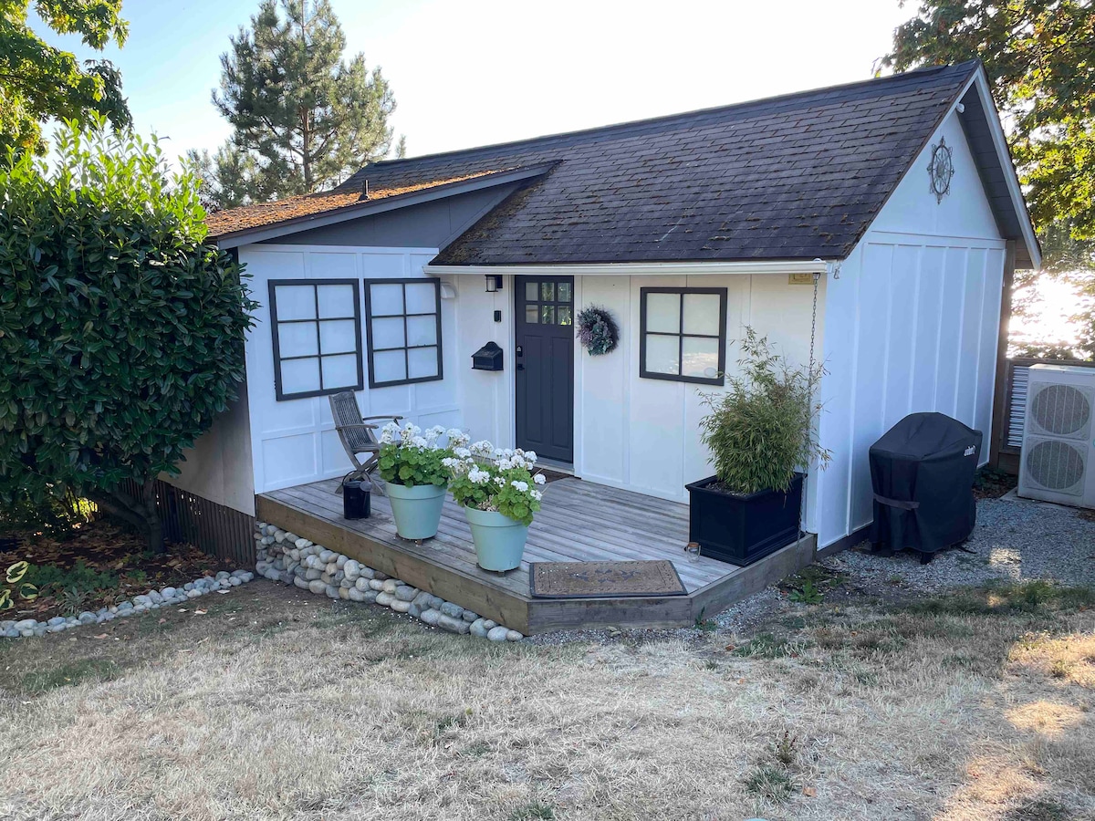 A charming cottage exterior is presented, featuring a small porch with potted plants and a black outdoor chair. Light-colored siding contrasts with the dark door and window frames. The landscape is maintained with grassy areas and stone borders, surrounded by trees and shrubs.