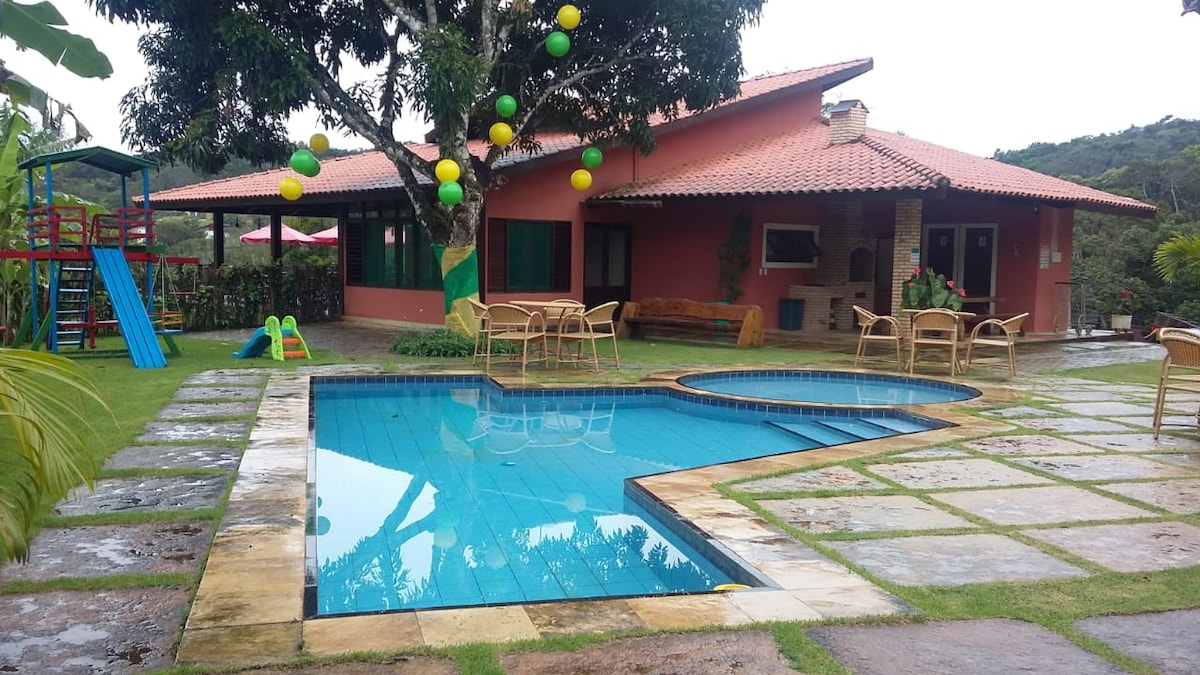 An outdoor area features a sparkling pool with a shallow section, surrounded by a patio with classic wooden chairs. The lush green lawn contains a children's playset and is complemented by vibrant balloons hanging in the nearby trees.
