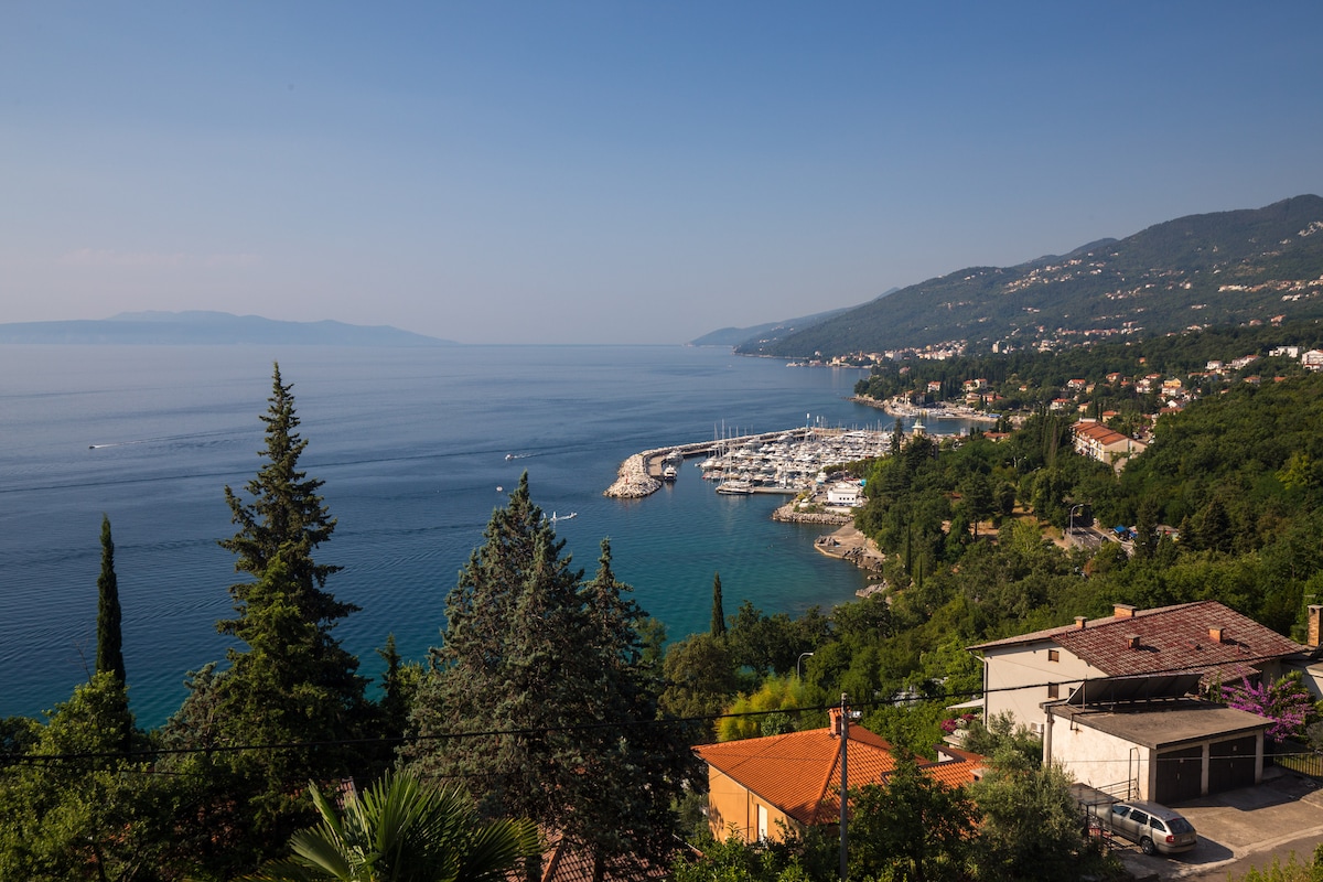 A serene coastal view showcases calm blue waters of the Kvarner Bay, dotted with boats in a marina. Lush greenery and olive trees line the shore, while a quaint neighborhood of houses can be seen above the beach. Gentle hills rise in the background under a clear sky.