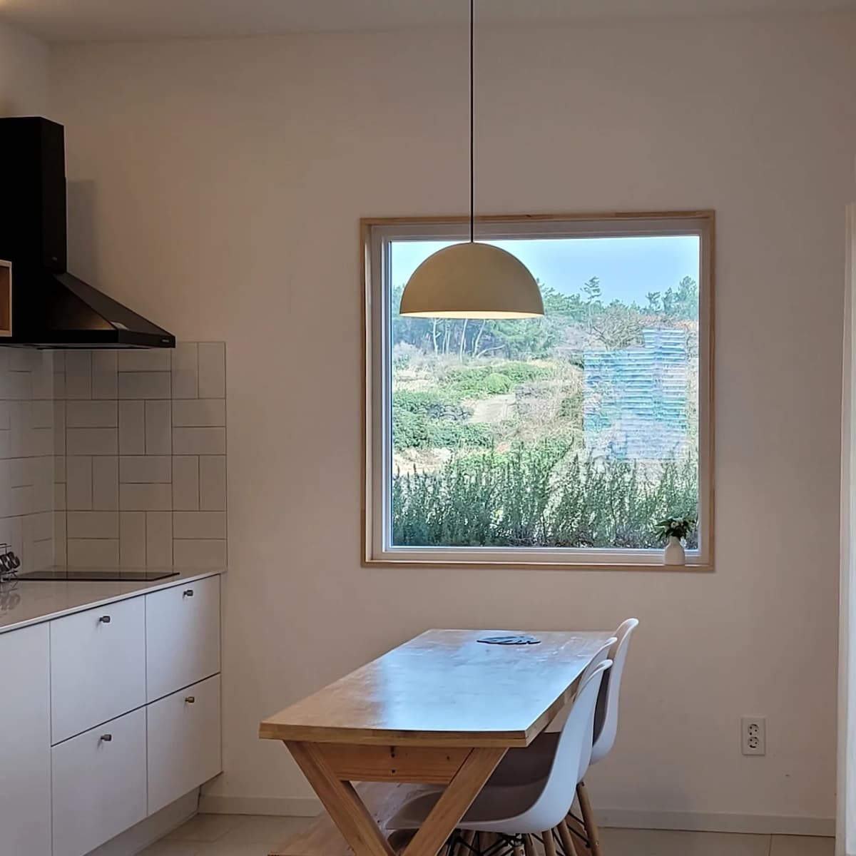 A clean, minimalist kitchen area is shown, featuring a wooden dining table with light-colored chairs. A window provides a view of the outdoor greenery, while a pendant light hangs above the table, adding functional illumination to the space.