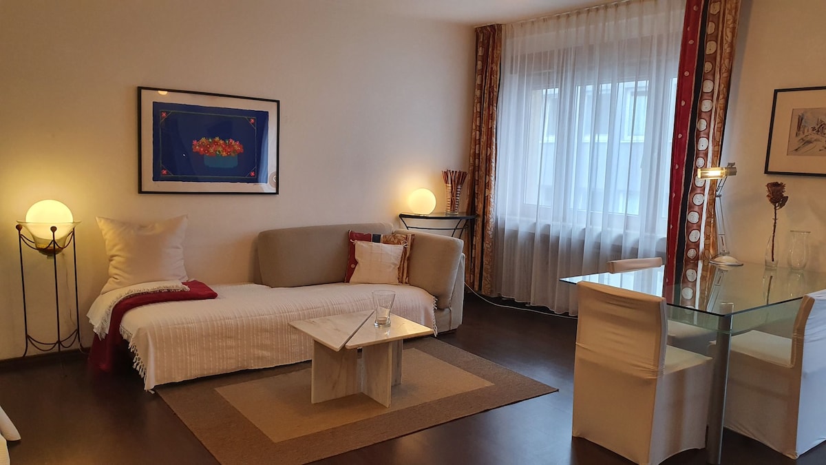 The living area features a light sofa and a cozy daybed, complemented by decorative pillows. A glass dining table is set with white chairs, while a piece of colorful artwork adorns the wall. Natural light filters through the sheer curtains, enhancing the inviting setting.