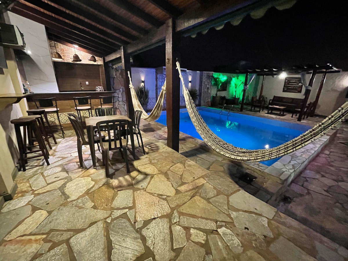 A spacious outdoor area is depicted, featuring a crystal-clear pool illuminated with soft lighting. Hammocks are suspended nearby, with a stone patio offering a dining set and bar. The roof showcases wooden beams, enhancing the ambiance for relaxation and leisure.