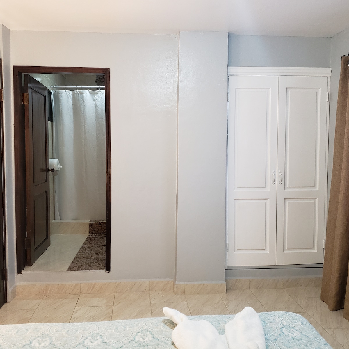 The room features a neutral color palette with smooth gray walls. A spacious closet with double doors is present, and a bathroom entry is visible in the background. The tiled floor complements the overall serene environment, while a hint of plush bedding is observable in the foreground.