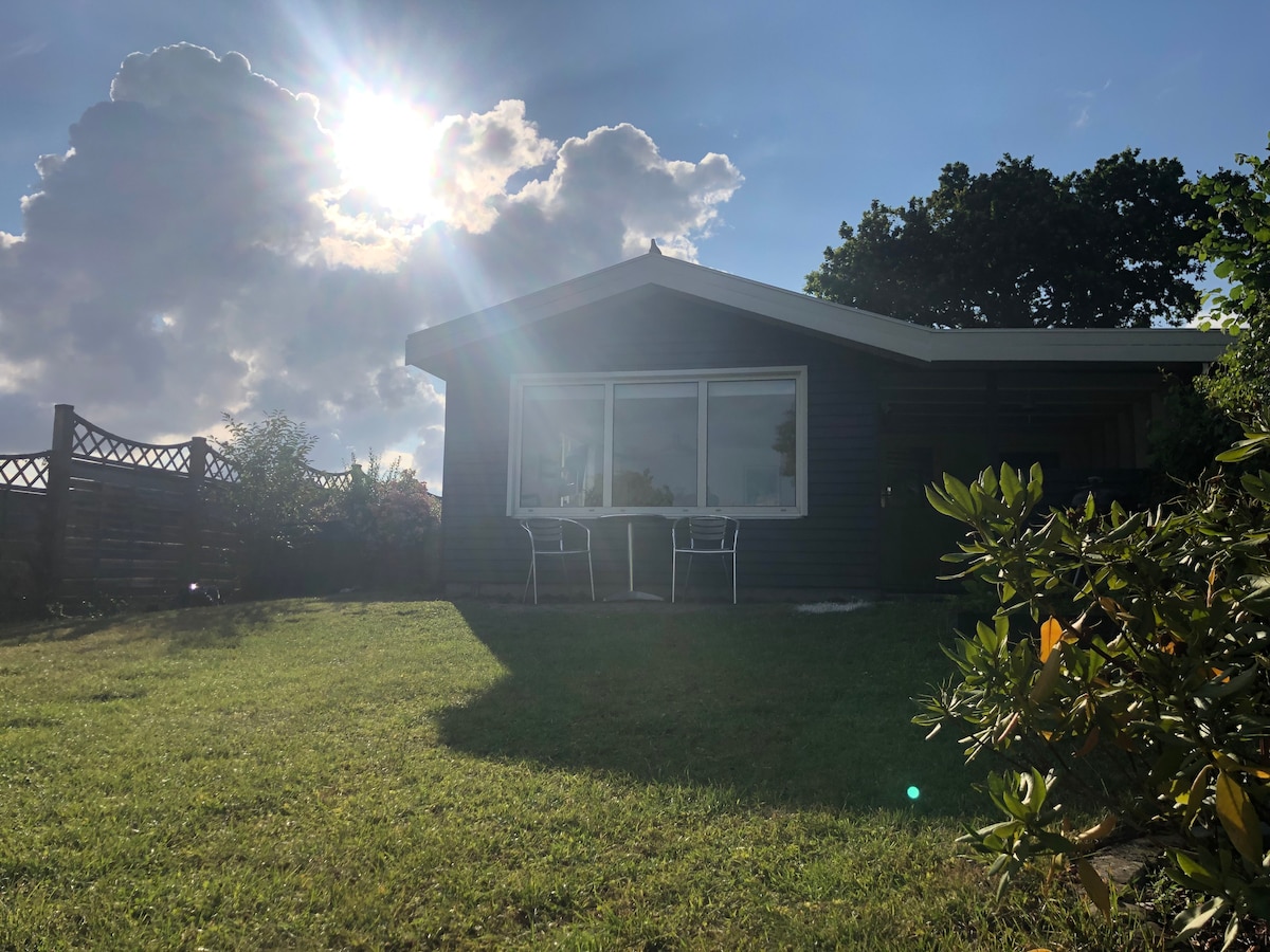 A charming summer house is pictured surrounded by greenery, with large windows reflecting sunlight. Two chairs are positioned outside, creating a serene setting. The sloping yard leads up to the house, providing a view of the sky partially covered by fluffy clouds.