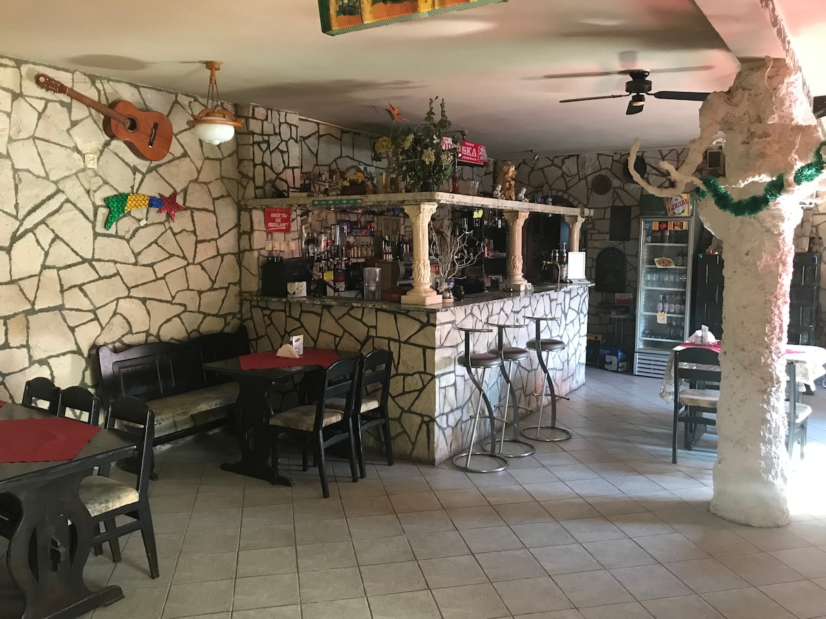 A unique bar area is presented with a stone wall design, featuring a wooden bar counter and seating arrangements. Tables with red tablecloths complement the rustic atmosphere, while decorative items, including a guitar and colorful wall art, add character to the space.