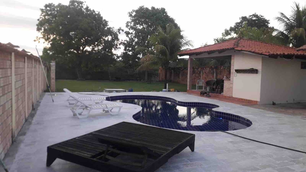 A view of a spacious outdoor area featuring a pool with an irregular shape, surrounded by well-maintained greenery. A wooden lounge chair is positioned beside the pool, with a covered seating area visible in the background, providing a sense of relaxation and comfort.