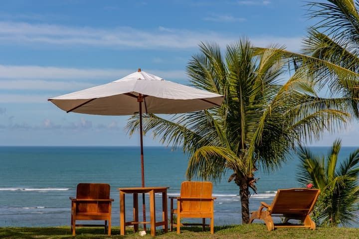 A serene outdoor setting showcases two wooden chairs and a table under a large umbrella, framed by palm trees. The ocean stretches beyond, reflecting the sky's blue tones, inviting relaxation amid a natural landscape.
