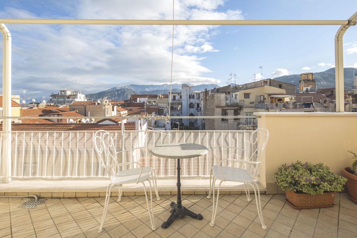 A spacious balcony features a round table accompanied by two white chairs, offering an unobstructed view of the rooftops of Sorrento. The surrounding buildings and distant mountains complement the scenic backdrop under a partly cloudy sky.