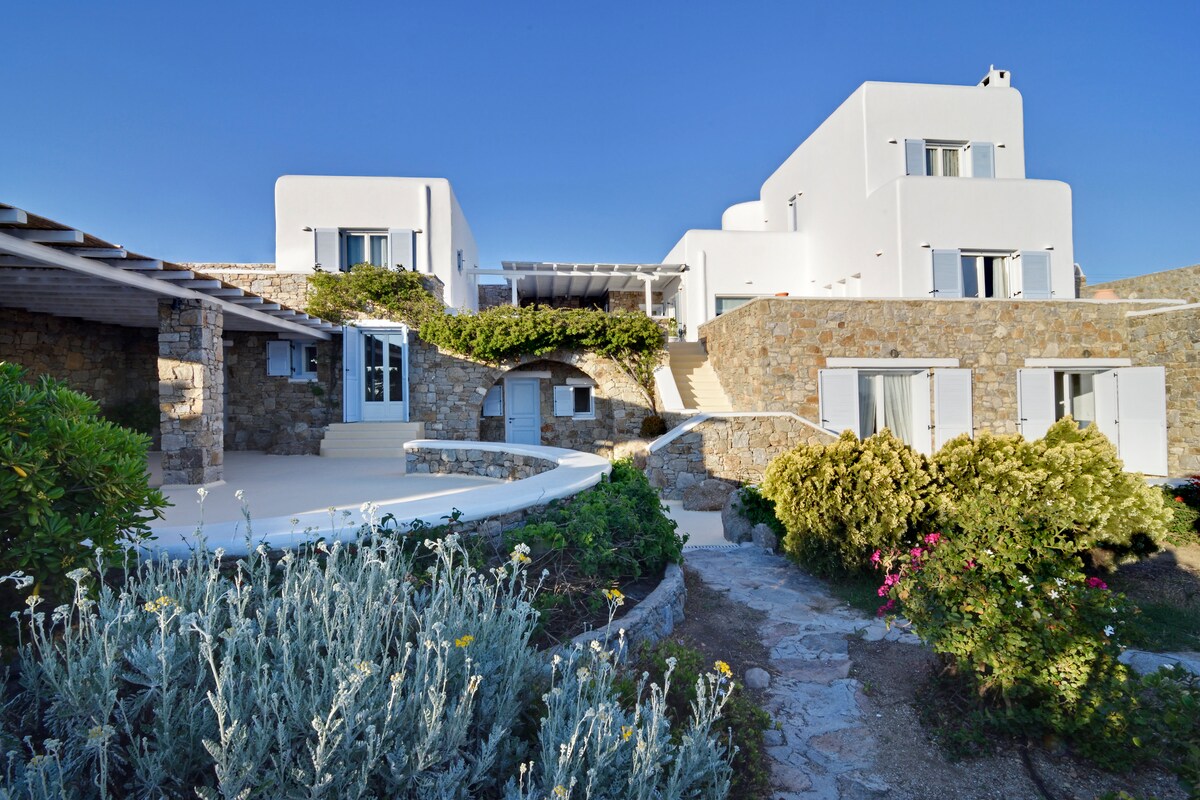 A modern white building is framed by lush greenery and stone pathways. The architecture features a combination of smooth surfaces and textured stone walls. Sunlight highlights the multiple levels of the property, creating inviting outdoor spaces with shaded areas for relaxation.