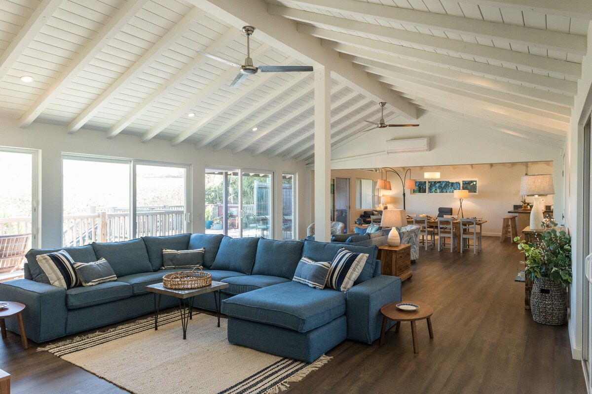 A spacious living area features a large sectional sofa in a calming blue hue, complemented by a light-colored area rug. Ample natural light floods in through expansive windows, offering views of the outdoor space. The open layout extends into a dining area with wooden furniture.