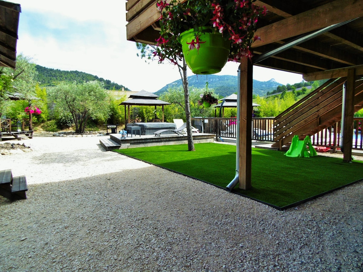 An outdoor space features a paved area leading to a grassy section, with ornamental plants in hanging pots. A jacuzzi and shaded seating can be seen, surrounded by lush greenery and hills in the background. The area is designed for relaxation and enjoyment.