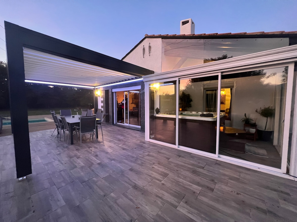 An expansive outdoor terrace is showcased, featuring a dining area with a large table and several chairs. A modern glass enclosure can be seen leading into the living space, highlighting a welcoming interior illuminated by soft lighting. The swimming pool is visible in the background.