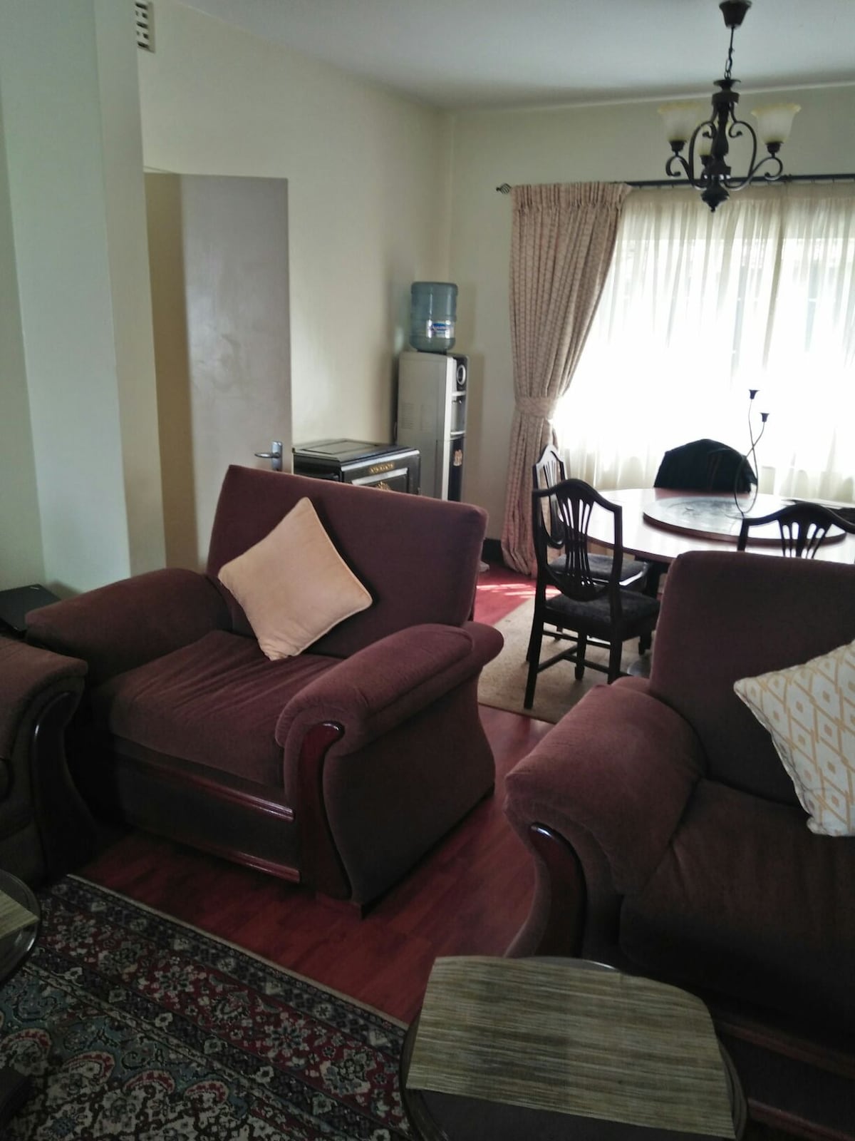 A comfortable living area is highlighted by two plush armchairs with decorative pillows. A round dining table is surrounded by four wooden chairs, while a water dispenser and a television are visible in the background. Natural light filters through sheer curtains.