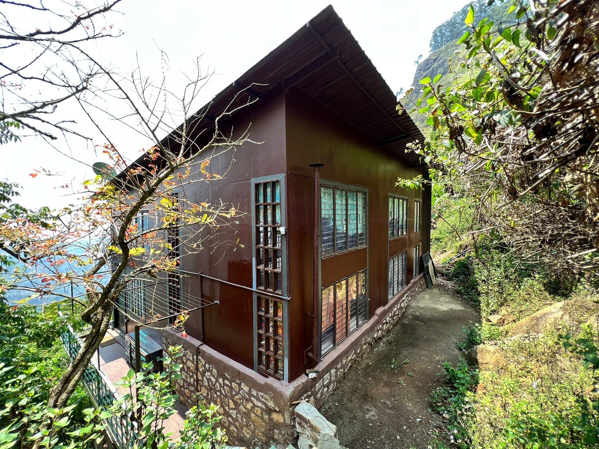 The exterior of the house is shown, featuring a sturdy structure with multiple windows framed in dark wood. A gravel path leads to the entrance, surrounded by lush greenery and trees. The building is nestled against the mountain backdrop, creating a harmonious presence in nature.