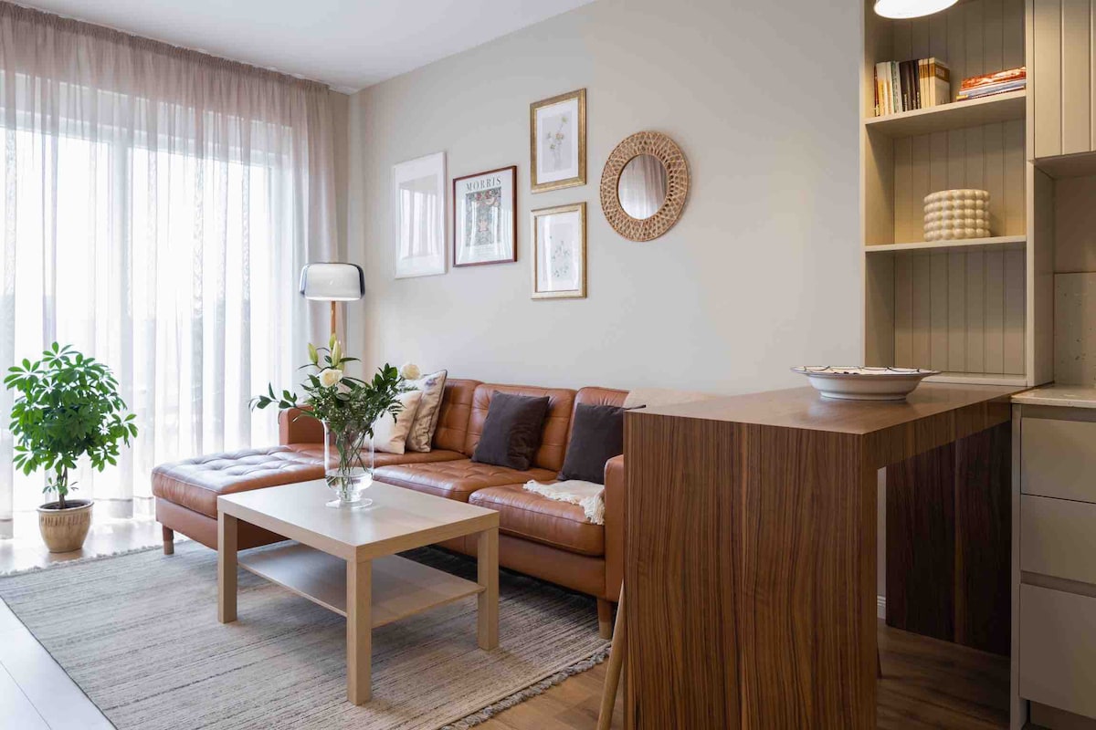 A spacious living area is featured with a leather sofa facing a coffee table. Natural light filters through sheer curtains, illuminating the room. Decorative wall art is displayed, and a small green plant adds a touch of nature beside the window.