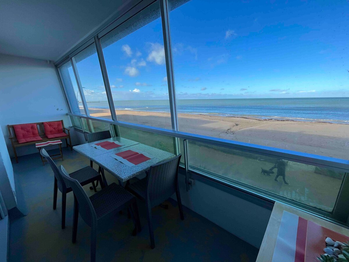 A bright loggia features large glass windows offering an unobstructed view of the beach and ocean. A table is set with a red and white table runner, surrounded by woven chairs. Plush seating with cushions is positioned in the corner, inviting relaxation.