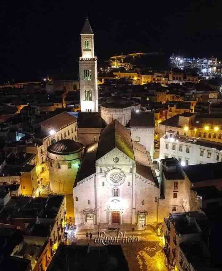 An aerial view presents a historic church illuminated at night, with its distinct bell tower rising prominently. Surrounding buildings create a dense, charming urban landscape, glimmering warm lights reflecting the town's vibrant atmosphere.