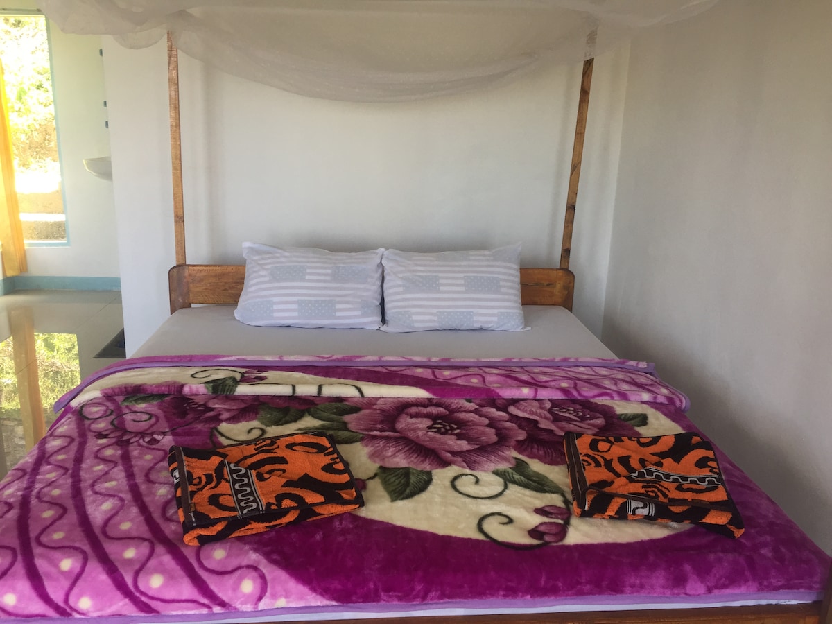 A spacious bed with a decorative purple and white blanket is centered in the room. Two patterned towels are neatly placed at the foot of the bed. Light streams through a nearby window, illuminating the simple, clean design of the interior.