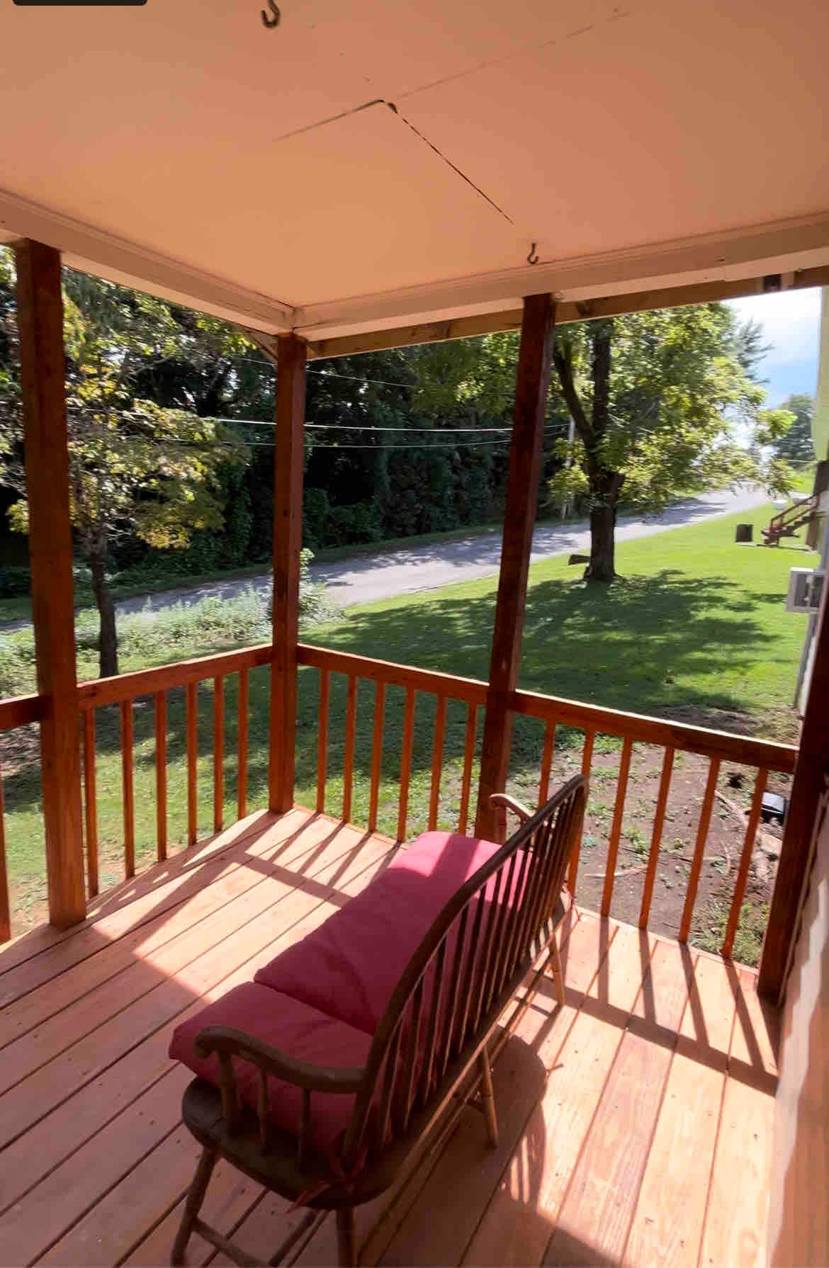 A covered porch is depicted, featuring a wooden railing and a single rocking chair with a burgundy cushion. Sunlight filters through the surrounding trees, and a view of the grassy yard and distant road is visible, creating a serene outdoor space.