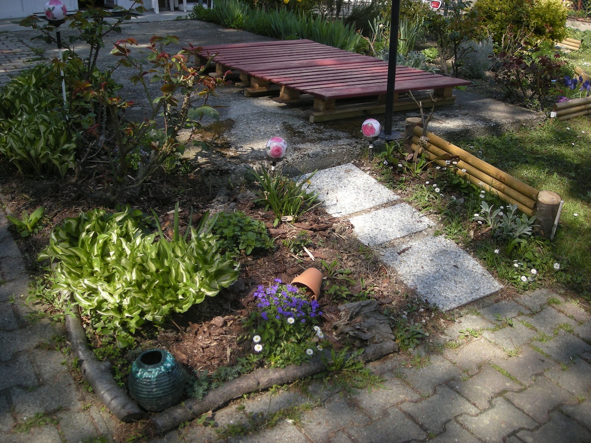 A well-maintained garden area features a mix of flowering plants and greenery, with a path leading through it. A wooden platform is visible in the background, surrounded by various decorative elements such as lanterns and a clay pot. The scene is brightened by seasonal blooms.