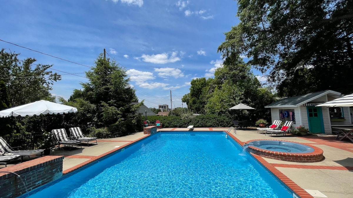 A large swimming pool glimmers under a clear blue sky, surrounded by lounge chairs. A hot tub is situated beside the pool, and shaded seating areas with umbrellas can be seen. Lush greenery adds to the tranquil outdoor space.