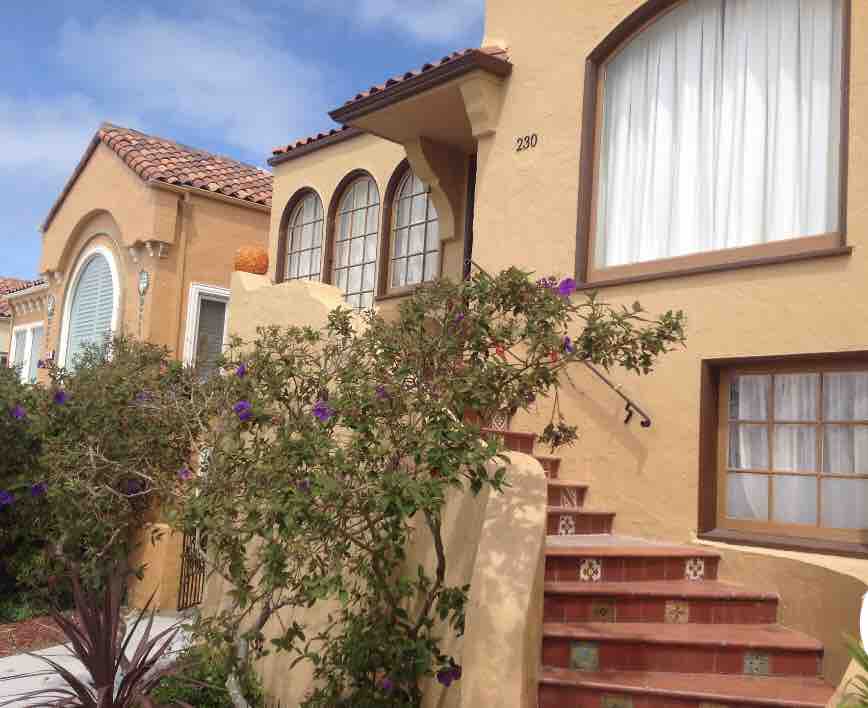 The exterior of a charming home is displayed, featuring a tiled staircase adorned with decorative details. Lush green plants and purple flowers frame the entrance, enhancing the welcoming atmosphere. Three arched windows offer glimpses of the light-filled interior behind soft curtains.