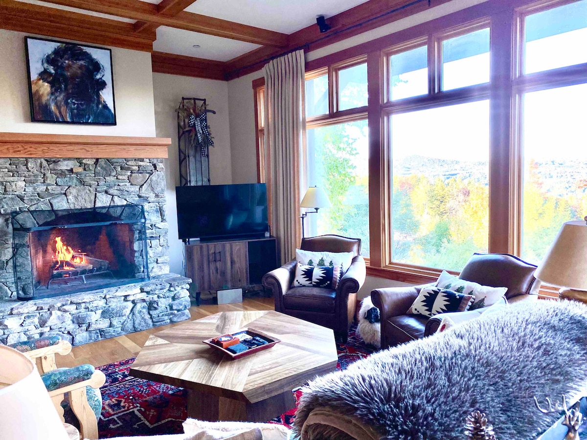 A cozy living area features a stone fireplace with a roaring fire, surrounded by large windows showcasing natural light and scenic views. Comfortable seating includes leather chairs and a patterned rug beneath a wooden coffee table, providing a welcoming space for relaxation.