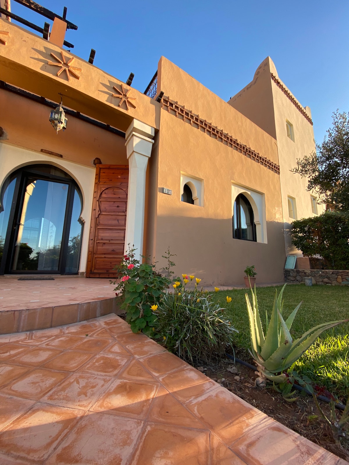 The entrance of a villa is presented, featuring a wooden door framed by arched windows. The exterior is adorned with warm earth-toned walls and decorative elements. Colorful flowers and green plants accentuate the pathway, leading to a well-maintained lawn under a clear blue sky.