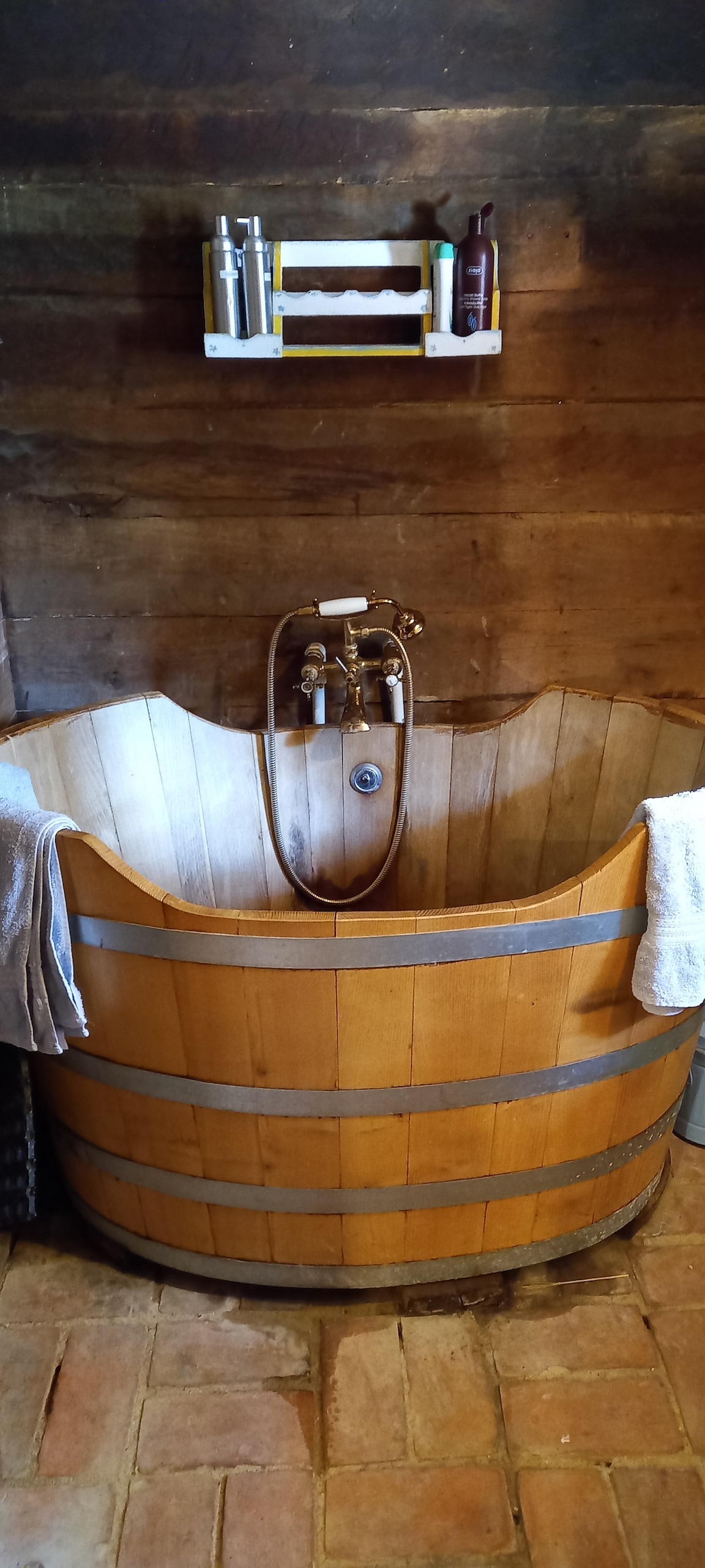 A handcrafted wooden bathtub is featured, showcasing a natural finish with metal bands. Above the tub, bathroom supplies are organized on a wall-mounted shelf. Soft, white towels are placed on the rim, complementing the rustic wooden wall and tiled floor.