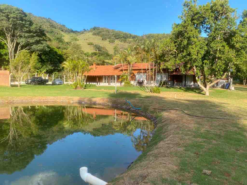 A serene landscape is displayed, featuring a tranquil pond reflecting the surrounding greenery and the house. Lush trees frame the property, and hills can be seen in the background, contributing to the peaceful atmosphere of the site.