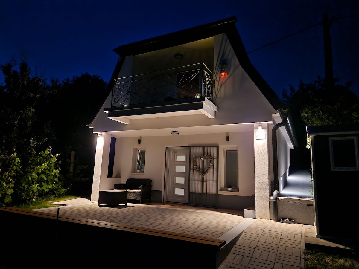 The exterior of a two-story house is illuminated at night, highlighting modern architectural features. A covered terrace extends at the front, furnished with seating. Soft lighting enhances the entrance, where a stylish door and large windows promote a welcoming appearance.